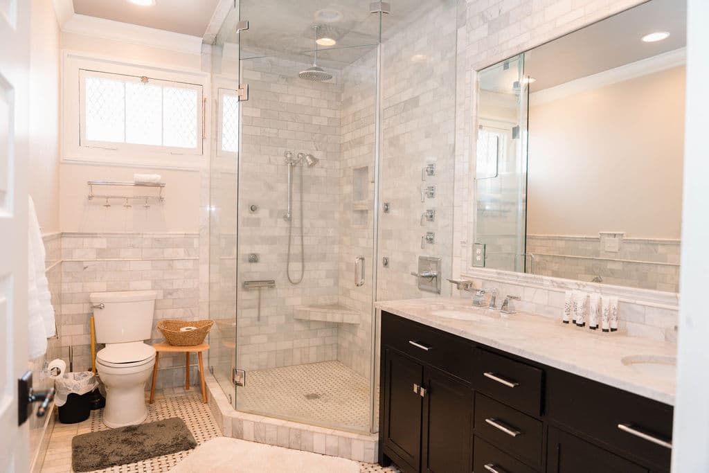 Full view of a luxury bathroom featuring a dark wood double vanity, a large glass-enclosed walk-in shower with marble tiles and multiple body jets, and a toilet situated beneath a small high window.