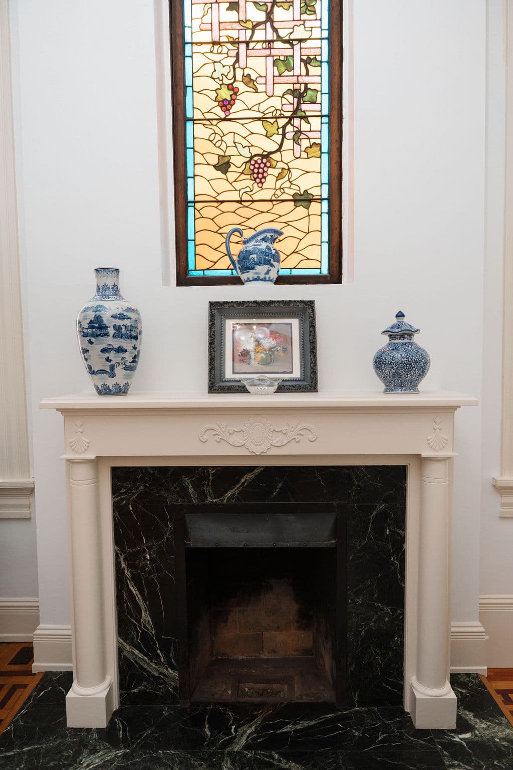 Classic fireplace design highlighting the dark marble hearth, ornate white mantel, and original stained glass accent window.