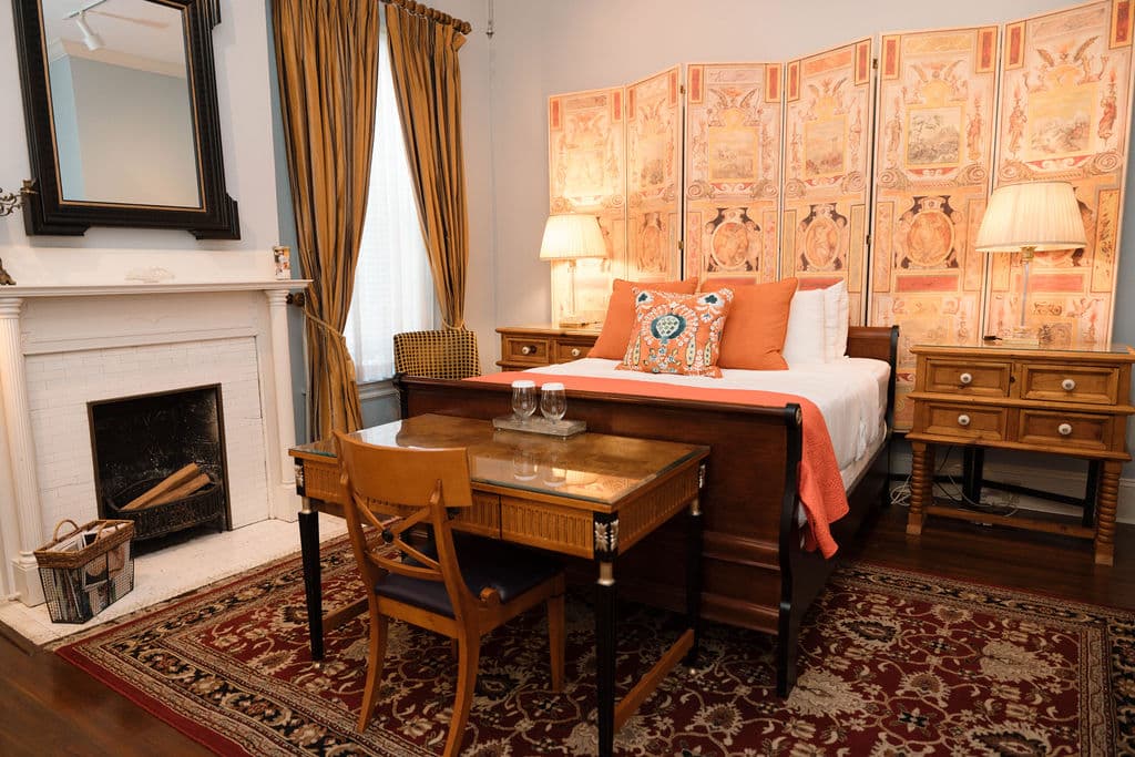 Bedroom suite featuring a wooden sleigh bed with orange accents, a decorative folding screen, antique desk, and white fireplace.