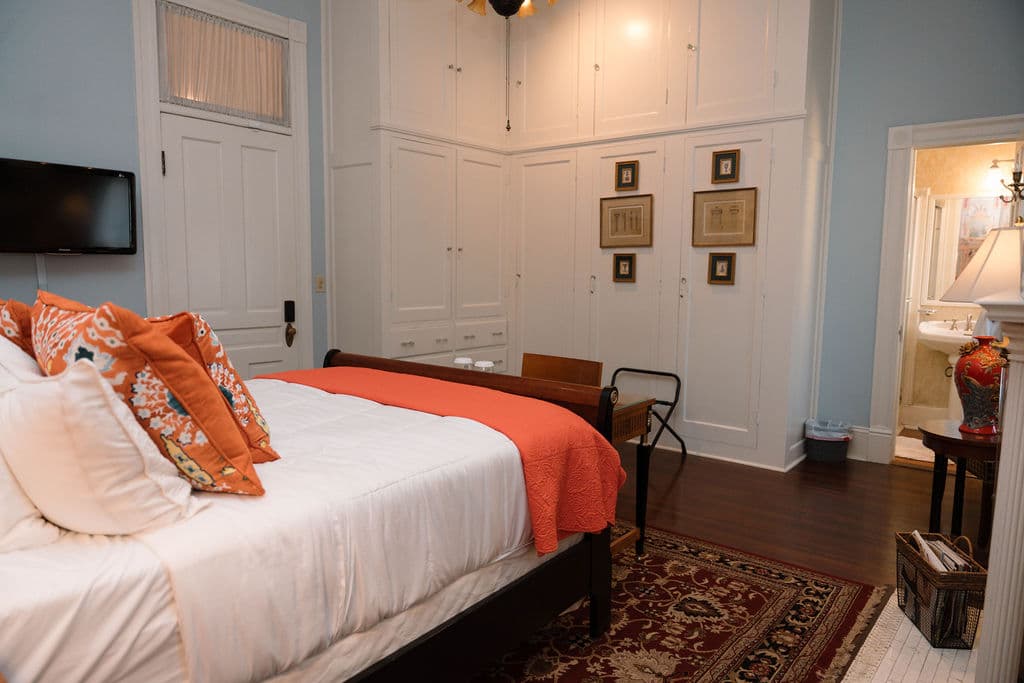 Bedroom suite featuring a sleigh bed with orange accents, a wall-mounted TV, floor-to-ceiling white built-in closets, and a view into the ensuite bathroom.
