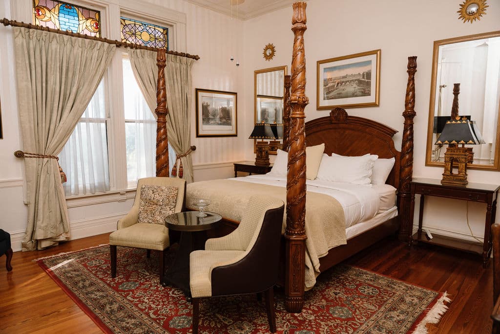 Elegant bedroom suite with antique-style furniture, stained glass window transoms, and a large wooden four-poster bed.