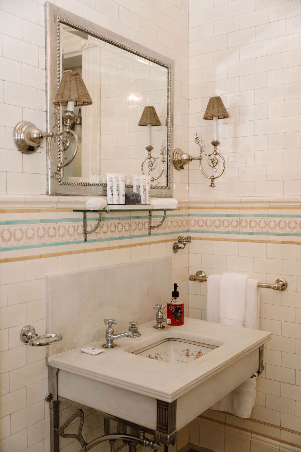 Detail of vintage-style bathroom fixtures including a console sink, chrome faucets, and wall-mounted lighting against a tiled wall with a colorful accent stripe.