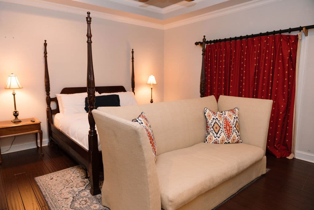 Traditional bedroom design highlighting a classic four-poster bed frame, a cozy seating area with a high-back beige settee, and dramatic red window treatments.