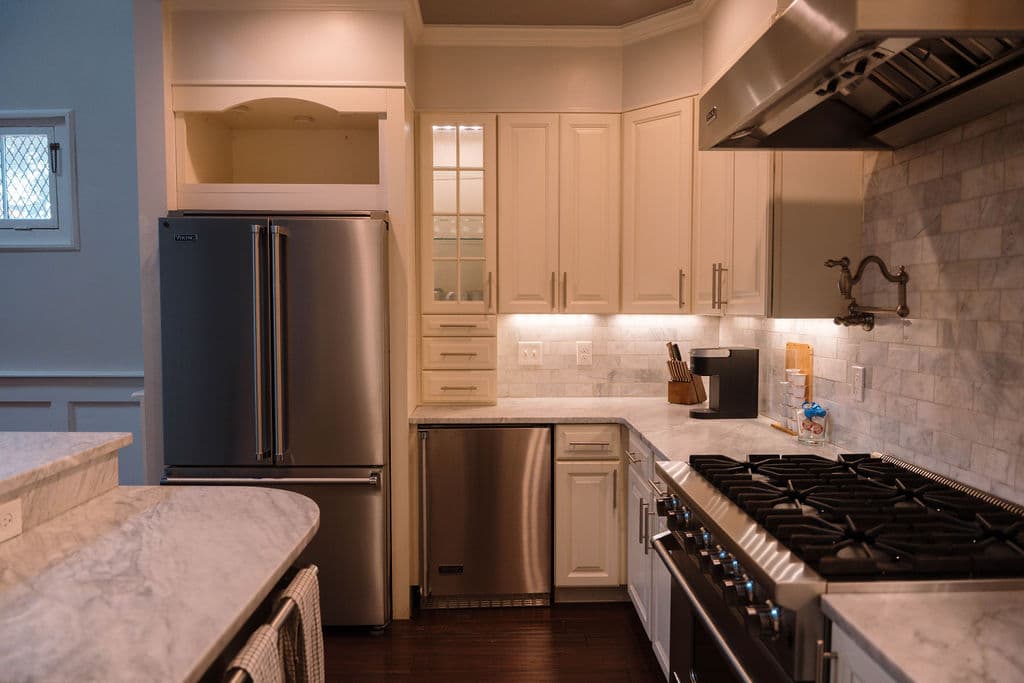 Close-up view of a kitchen workspace featuring a stainless steel gas range with a wall-mounted pot filler faucet. The area includes a stainless steel dishwasher, a French-door refrigerator, and white cabinetry with under-cabinet lighting illuminating the marble backsplash.