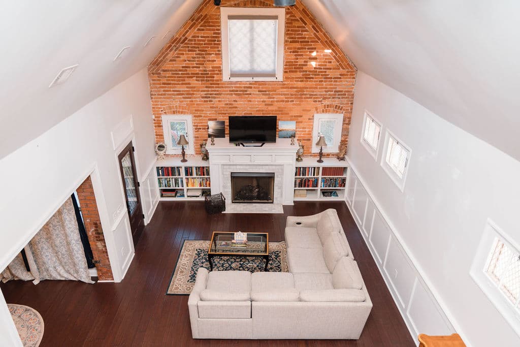 High-angle view of a spacious living room featuring a cathedral ceiling and a floor-to-ceiling exposed brick wall. The layout includes a large L-shaped beige sectional sofa facing a white fireplace mantel with built-in bookshelves. The room has dark hardwood floors and white wainscoting along the side walls.