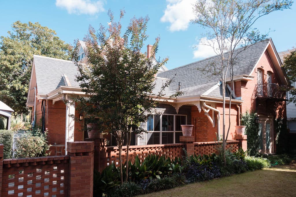 Exterior view of a historic red brick home featuring a steeply pitched roof, decorative wooden gable details, and large windows. The property is surrounded by a low brick privacy wall and lush green landscaping under a bright blue sky.