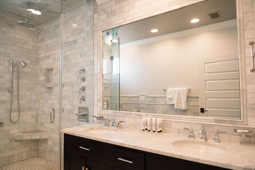 Modern bathroom featuring a dark wood double vanity with a marble countertop and a large mirror. To the left is a spacious glass-enclosed walk-in shower with gray tiled walls, a built-in bench, handheld showerhead, and body spray jets.