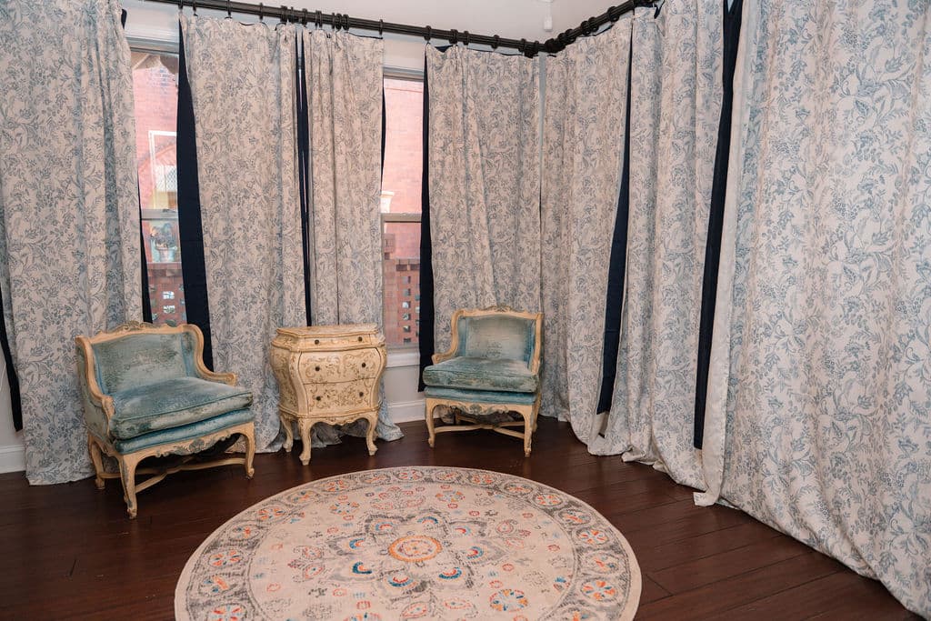 Corner seating area featuring two light blue French-style armchairs and a painted chest, surrounded by floor-to-ceiling blue and white floral curtains. A round floral rug sits on the dark hardwood floor.