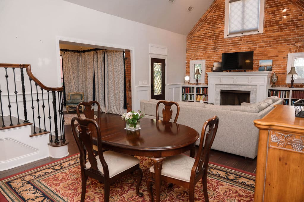 Open-concept dining area featuring a dark wood oval table and four chairs on a red patterned rug. To the left is a staircase with white steps and black metal railings. The space flows directly into a living room with a beige sectional sofa and an exposed brick wall. A doorway in the background offers a glimpse into a room with blue floral curtains.