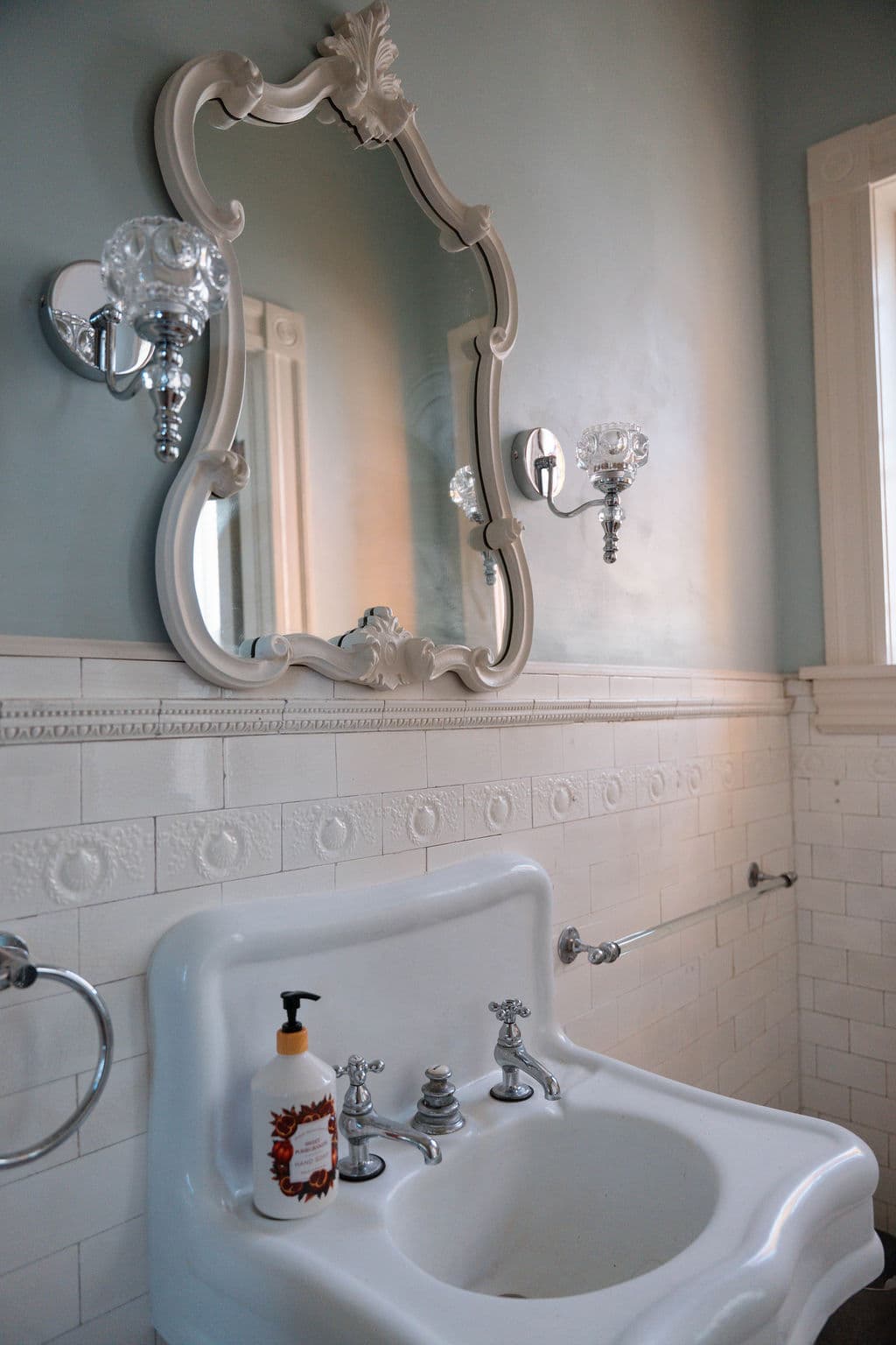 Romantic bathroom design highlighting a decorative mirror, soft blue walls, and classic white tiled wainscoting.