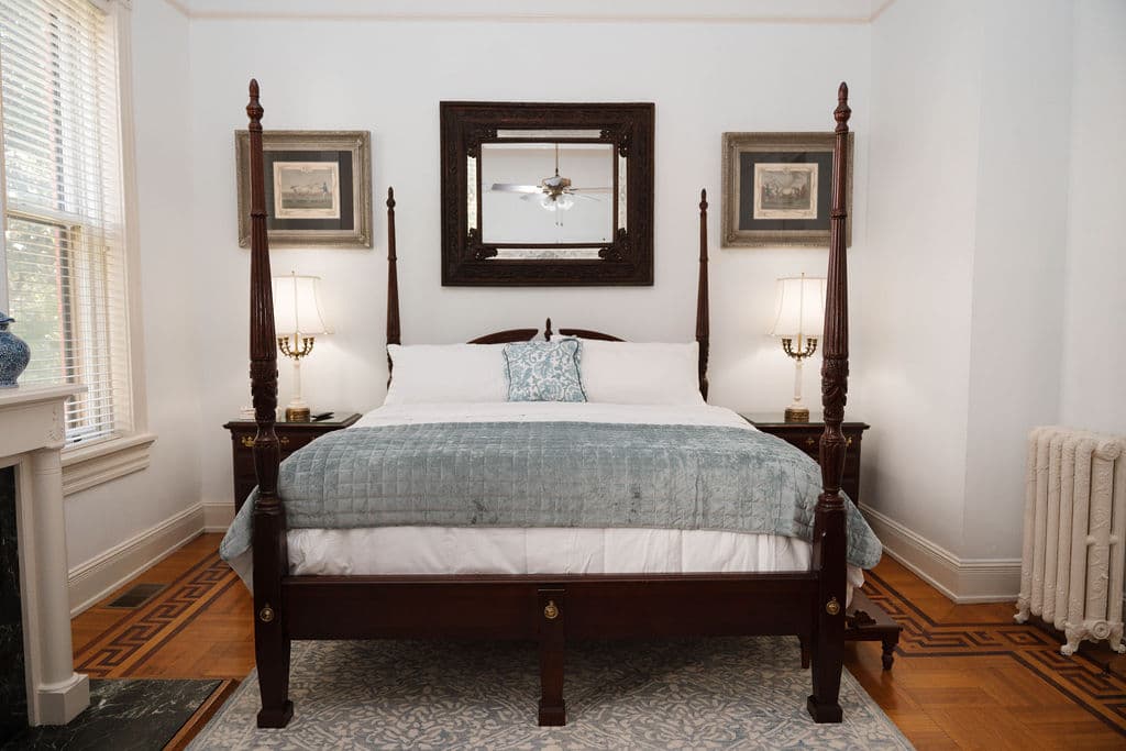 A bedroom featuring a dark wood four-poster bed with light blue bedding. A large dark-framed mirror hangs above the headboard, flanked by two framed artworks and bedside lamps. The room features original parquet hardwood flooring and a white radiator.