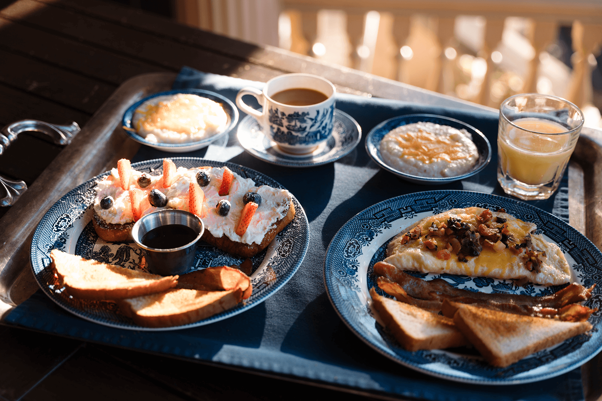 A breakfast tray with two plates, one with fruit-topped toast and syrup, the other with an omelette, toast, and bacon. Includes coffee, juice, and oatmeal.