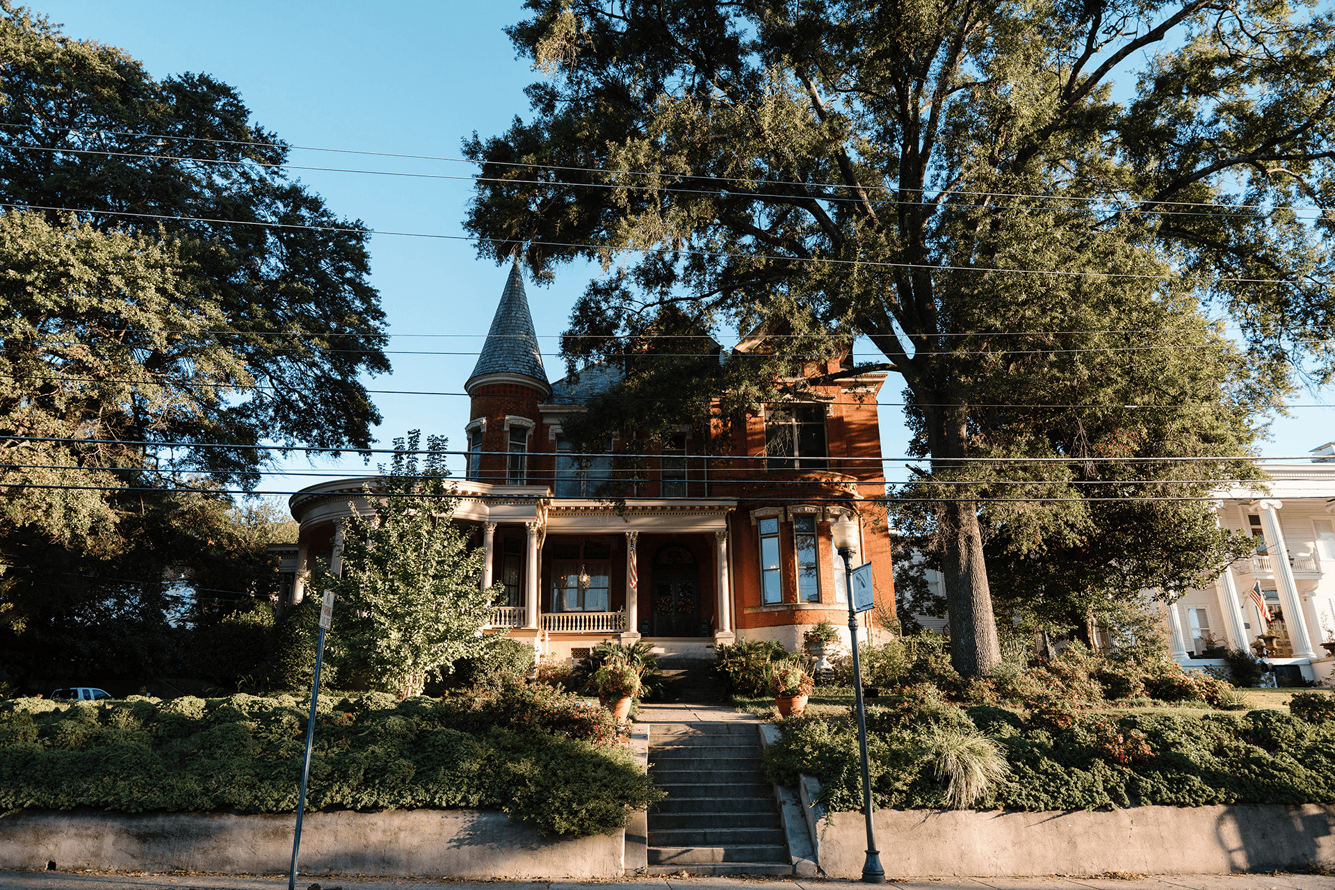 A historic two-story brick house with a tower, surrounded by lush greenery and trees.
