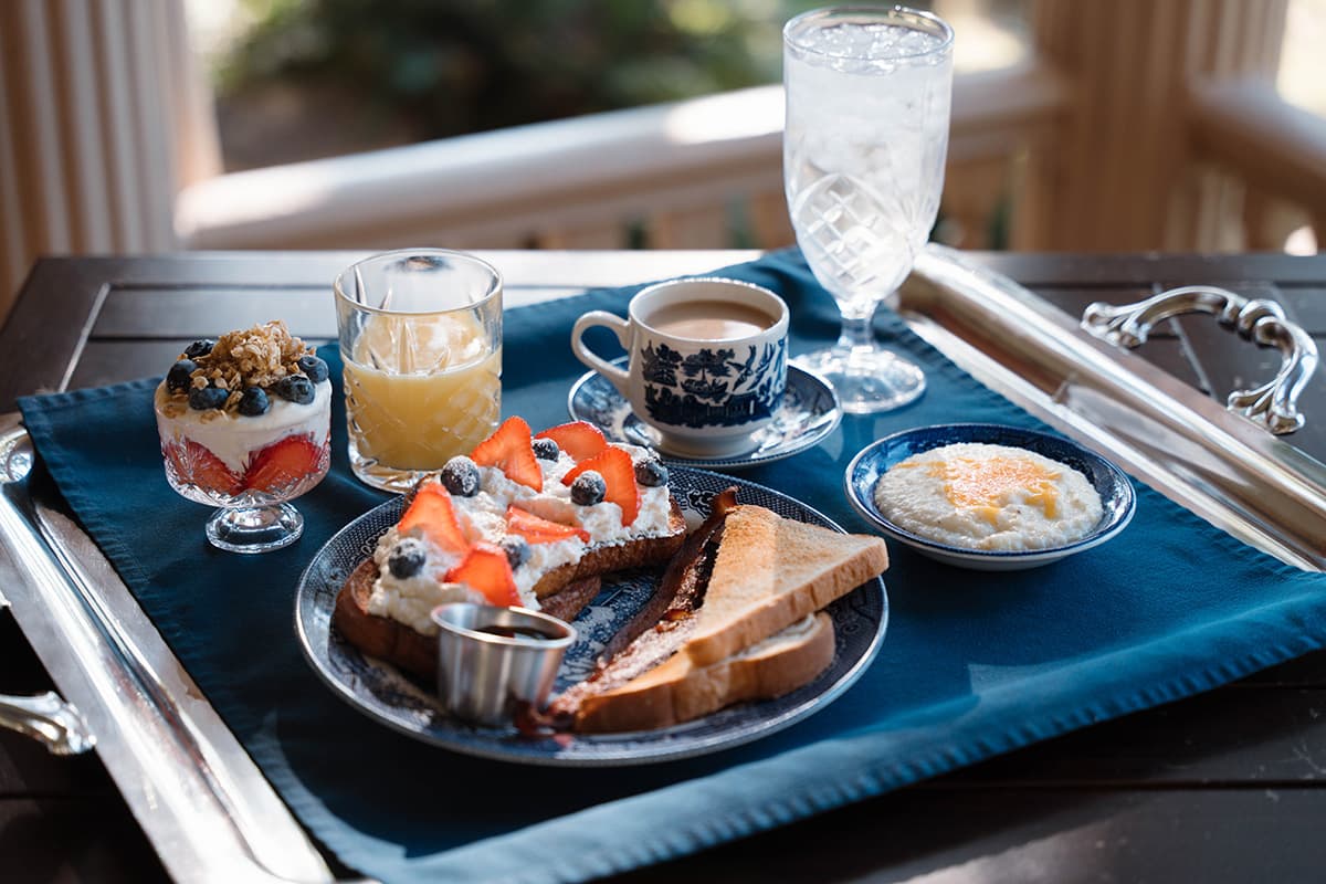 A breakfast tray featuring toast with fruit, a yogurt parfait, coffee, juice, and water, set on a blue cloth.