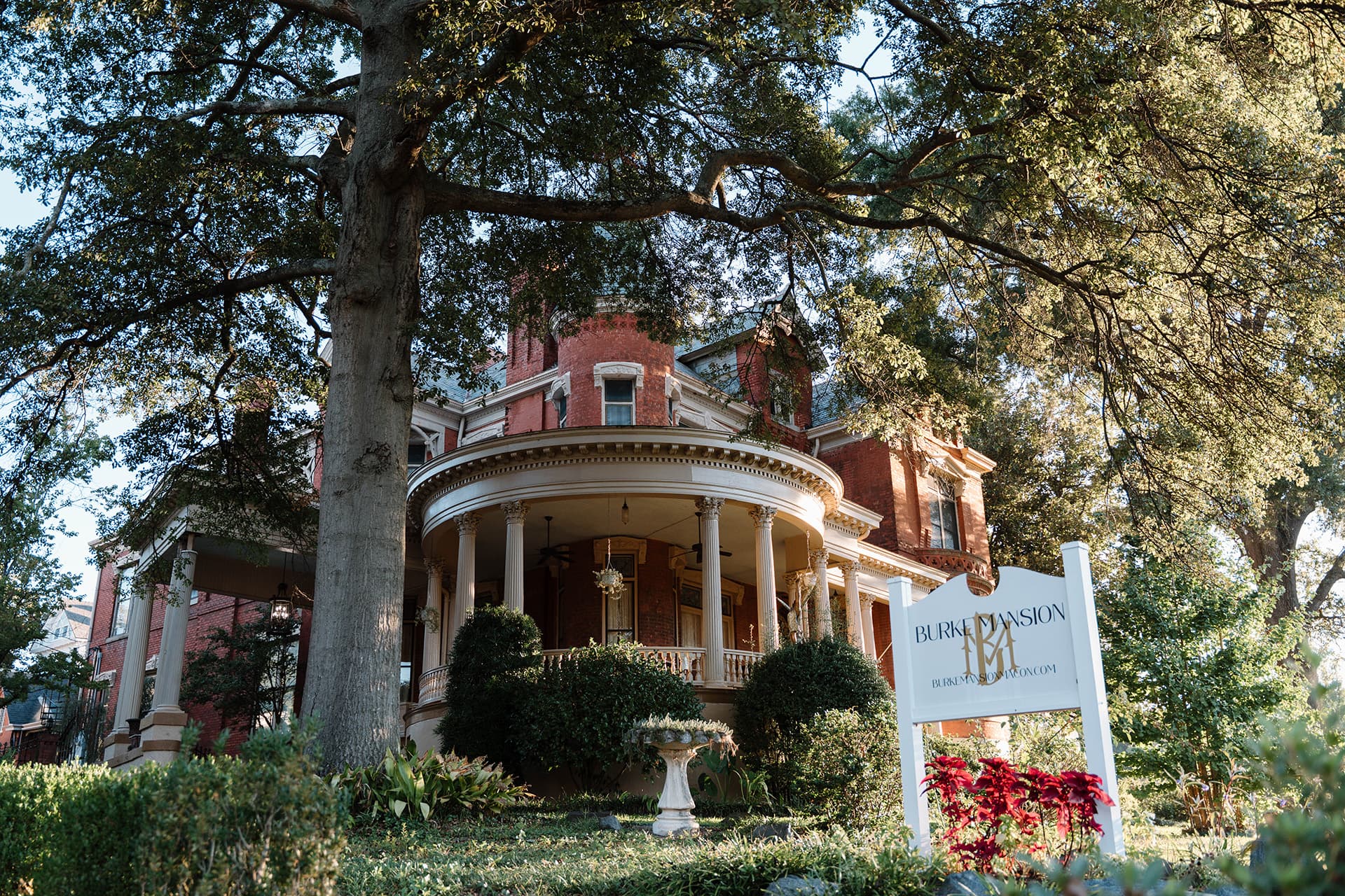 A historic red mansion with a wraparound porch, surrounded by greenery and a welcoming sign.