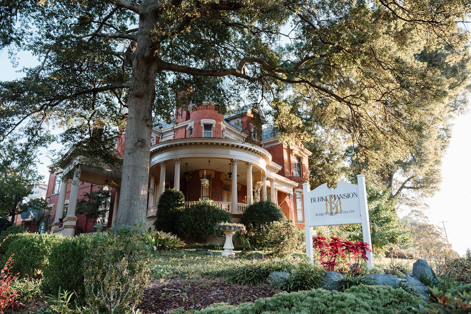 A historic red mansion with a wraparound porch, surrounded by greenery and a welcoming sign.